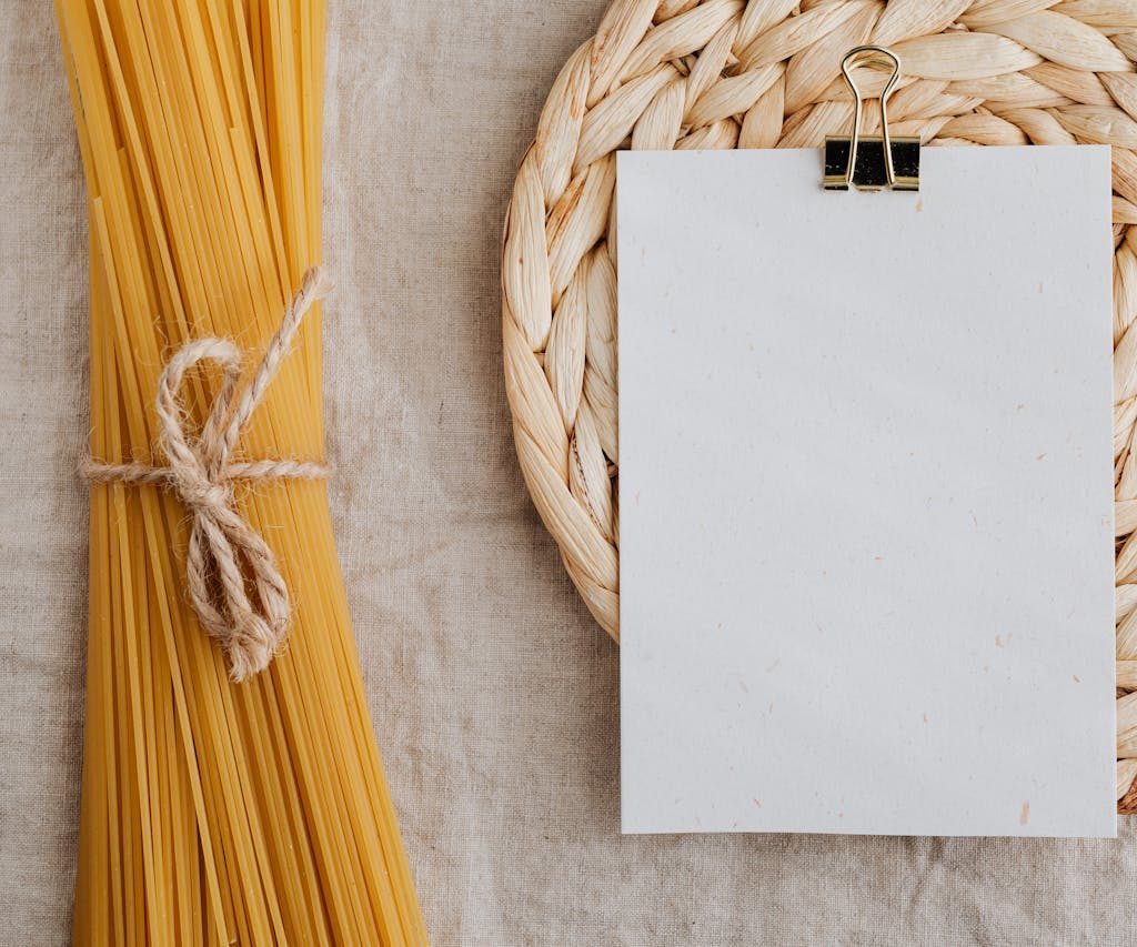 From above of blank paper placed on wicker table mat next to spaghetti tied with bow arranged on table covered with linen tablecloth suitable for recipe or ingredient listing